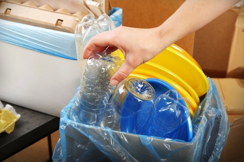 A person’s hand is seen placing a clear plastic water bottle into a blue trash bag lined with a transparent plastic sheet. Inside the bag, there are several bright yellow and blue plastic buckets stacked together, with their handles visible. The background includes a portion of a wooden surface or furniture, suggesting an indoor or garage setting. The image highlights the process of disposing of household waste, specifically recyclable plastic items, which is relevant to rubbish removal services such as those provided by Garden Clearance St Johns Wood. The scene emphasizes organized waste segregation with focus on plastic containers, suitable for outdoor waste removal or clearance in the St Johns Wood area, known for residential properties and garden maintenance needs. The lighting is natural or warm, illuminating the plastic items clearly, reflecting typical domestic waste disposal activities commonly encountered in urban environments like St Johns Wood.