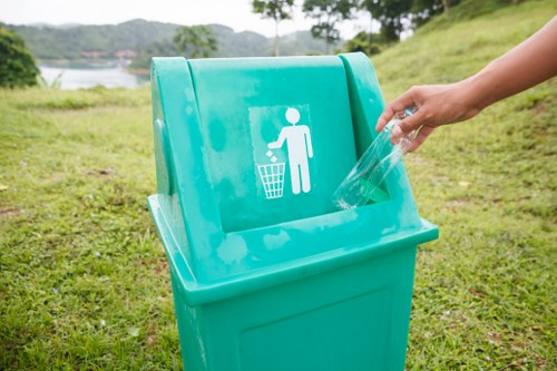 A close-up view of a bright blue urban recycling bin situated outdoors on a grassy area, with a white graphic depicting a person throwing waste into a bin displayed on the front. A person's hand, visible on the right side of the image, is placing a transparent plastic bottle into the bin. The background shows a natural, green landscape with gentle slopes, trees, and distant water, indicating an open outdoor environment typical of a park or outdoor recreational space near St Johns Wood. The bin's surface appears clean and smooth, with the environment suggesting a clean-up or rubbish disposal activity. This scene captures the practice of waste management and recycling in a local outdoor setting, relevant to rubbish removal services in the area.