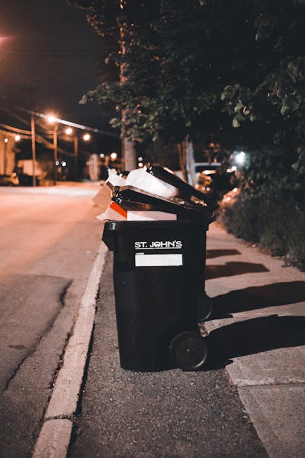 A black wheeled rubbish bin labeled 'St. John's' stands on a sidewalk beside a street at night. The bin's lid is open, revealing various discarded paper and cardboard materials. The bin is positioned 